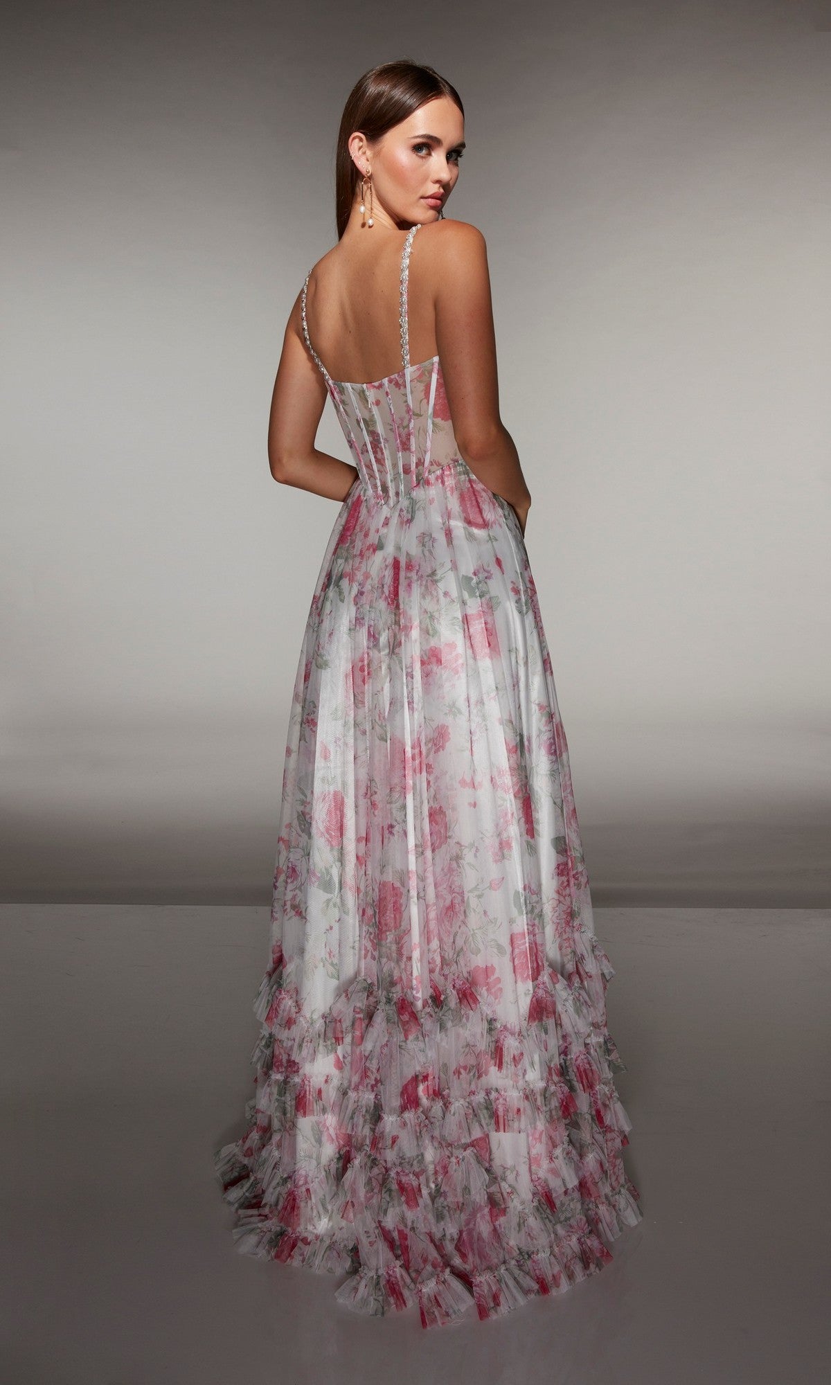 A woman wearing the Alyce 62202 long prom dress, featuring a sleeveless floral print, layered ruffled skirt, and sheer corset bodice, stands with her back to the camera.